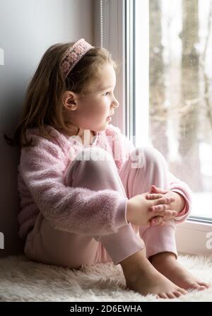 Kid sitting on windowsill at home looks out window to snow, portrait of pretty little girl on fur rug on room sill in winter. Adorable pensive child i Stock Photo