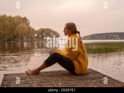 A barefoot girl with her hair in braids sits on a wooden bridge by the lake against the sunset. The girl is holding a Cup with a drink.  Stock Photo