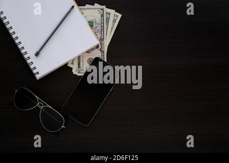 Dark wooden desk with objects. Top view of a notebook, money, cell, phone, pen and sunglasses. Stock Photo