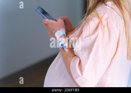 Pregnant Caucasian woman with drip infusion needle in her arm, using smartphone. Giving birth at hospital. Stock Photo