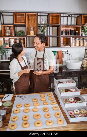 Asian couple smiles while using their cellphone after making donuts together in the kitchen Stock Photo