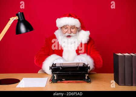 Photo of amazed santa claus sit table desk work computer impressed many  wish gift list letters in house idnoors with evergreen christmas x-mas tree  ornament decoration wear cap headwear Stock Photo