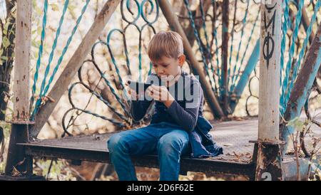 Focused boy using smartphone in park. Serious concentrated school aged boy in casual wear sitting on bench and playing game on mobile phone while Stock Photo