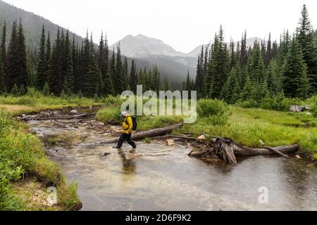 Side view of man hiking in mountains and crossing river on overcast day in British Columbia Stock Photo