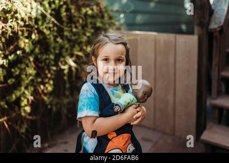 Little girl carrying babydoll in baby carrier outside in yard Stock Photo