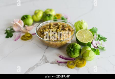 Bowl of tomatillo salsa with it's ingredients on white quartz counter. Stock Photo