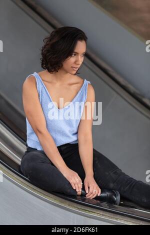 young brunette latina woman with short hair with curls, sitting and distracted on a handrail of electric bleachers, wearing blue blouse, black pants Stock Photo