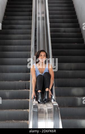 cheerful young brunette latina girl with short hair with curls, sitting on the handrail of electric bleachers, wearing blue blouse, black pants Stock Photo
