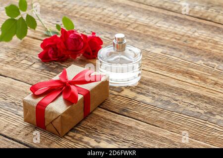 Gift box wrapped with a ribbon and a bottle of perfume on old wooden boards decorated with red roses. Top view. Stock Photo