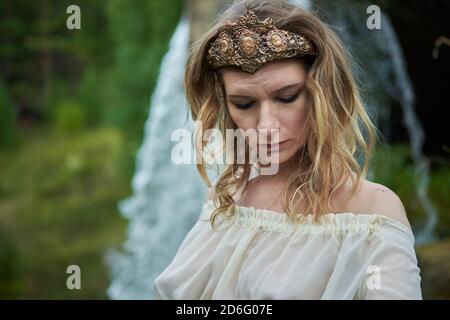 Young beautiful pregnant girl in a white dress and crown stands against the background of a waterfall  Stock Photo