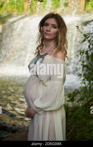 Young beautiful pregnant girl in a white dress and crown stands against the background of a waterfall  Stock Photo