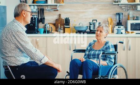 Retired invalid woman in wheelchair having a conversation with old elderly husband in kitchen. Old man talking with wife. Living with disabled person with walking disabilities Stock Photo