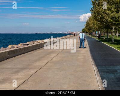 Istanbul / Turkey - 10.1.2020; An elderly man is walking with crutches on a park pavement surrounded by trees next to the sea on a sunny day. Stock Photo