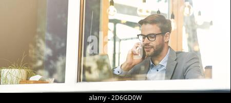 Young handsome businessman wearing eyeglasses talking by phone while working remotely in cafe Stock Photo