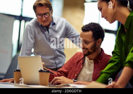 Successful project team. Group of focused multiracial business people looking at laptop screen and discussing work or project results while standing in the modern office or coworking space Stock Photo