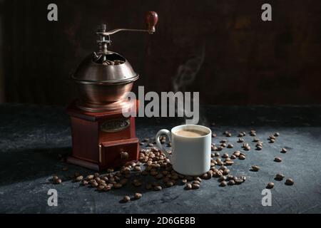 Manual wooden coffee grinder, white cup with the steaming hot drink and some roasted beans on a dark rustic background, copy space, selected focus, na Stock Photo
