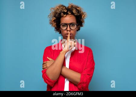 Pretty african woman makes shush, asks not be loud, has secret expression, presses index finger over lips Stock Photo