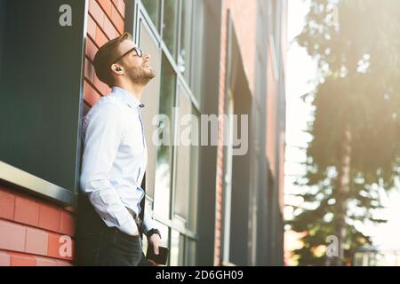 Side view of a young happy businessman listening to music with wireless earphones and mobile phone while leaning against brick wall outdoors Stock Photo