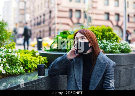Redhead woman in protective mask talking on a smartphone in downtown. Business during coronavirus concept. Stock Photo