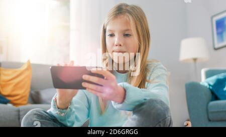 Portrait Shot of a Smart Cute Girl Sitting on a Carpet Playing in Video Game on His Smartphone, Holds and Uses Mobile Phone in Horizontal Landscape Stock Photo