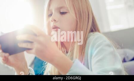 Close-Up Portrait of a Smart Cute Girl Sitting on a Carpet at Home Playing in Video Game on His Smartphone, Holds and Uses Mobile Phone in Horizontal Stock Photo