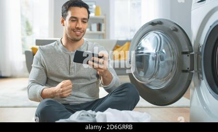 Handsome Smiling Young Man in Grey Jeans Sits in Front of a Washing Machine, Plays Games on His Smartphone. He win. He was Loading the Washer with Stock Photo