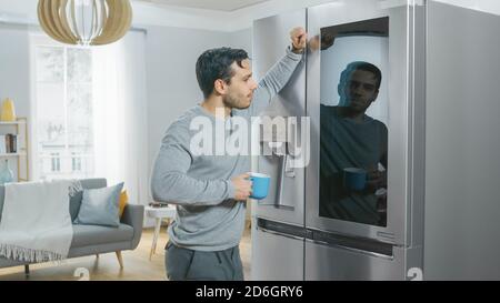 Handsome Young Man is Standing Next to a Refrigerator While Drinking His Morning Coffee. He is Checking a To Do List on a Smart Fridge at Home Stock Photo