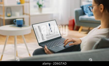 Young Talented Woman Architect is Sitting at Home on a Couch Working with a Laptop Computer under Futuristic Concept 3D Model of Modern Skyscraper. Stock Photo
