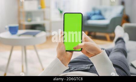 Woman at Home Lying on a Couch using Smartphone with Green Mock-up Screen, Doing Swiping, Scrolling Gestures. Girl Using Mobile Phone, Internet Social Stock Photo