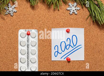 White pills, white sticky note with 2020 pinned on notice board which is decorated with pine twigs and snowflakes at the top. Healthcare, christmas, n Stock Photo
