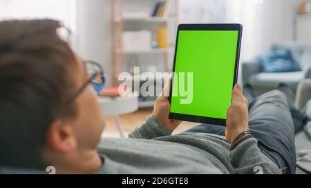 Young Man at Home Laying on a Couch Using with Green Mock-up Screen Tablet Computer in Vertical Portrait Mode. Man Using Touchscreen Device, Browsing Stock Photo
