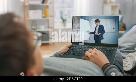 Man at Home Laying on a Couch Using Laptop Computer for Video Call to His Teacher. Screen Shows Online Lecture with Teacher Explaining Math Subject.  Stock Photo