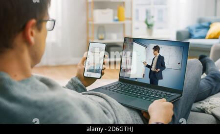 Young Man at Home Works on a Laptop Computer sing Laptop Computer for Video Call to His Business Analyst, while Holding Smartphone, Showing Statistics Stock Photo
