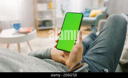 Man at Home Lying on a Couch using Smartphone with Green Mock-up Screen. Over the Shoulder Camera Shot. Stock Photo