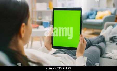 Young Woman at Home Resting on a Couch Using with Green Mock-up Screen Tablet Computer in Vertical Portrait Mode. Woman Using Tablet Device, Browsing Stock Photo