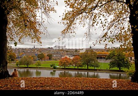 Inverleith Park, Edinburgh, Scotland, UK. 17 October 2020. On an overcast day trees beginning to lose their Autumn colours in Inverleith with the city centre skyline in the background.One lady sits under a tree admiring the view over the pond. Credit: Arch White/Alamy Live News Stock Photo