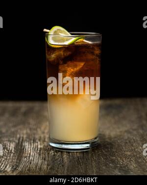 Dark and Stormy cocktail in high ball glass with ice and a lime garnish on an old wooden table with a black background Stock Photo
