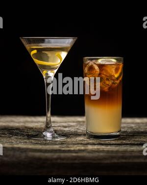 Dark and Stormy cocktail in high ball glass with ice and a lemon garnish with a Vodka Martini on an old wooden table with a black background Stock Photo