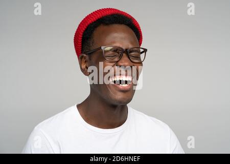Laughing positive young Afro American man wear red hat in good mood, poses at camera with closed eyes. Overjoyed Black male in spectacles shows her br Stock Photo