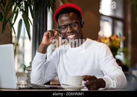 Happy millennial Black man in headphones wear red hat, enjoying watching educational webinar on laptop, remotely online work in cafe, holding cup of c Stock Photo