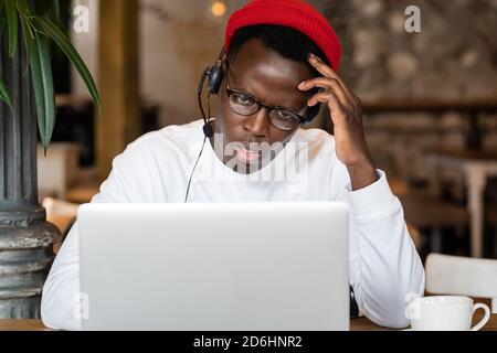 Thoughtful millennial Black man in headphones wear red hat, watching educational webinar on laptop, remotely online work in cafe. Afro-American male l Stock Photo