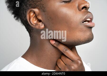 Closeup of afro man touches fingers of sore throat, isolated on gray background. Thyroid gland, painful swallowing, tonsillitis, laryngeal swelling co Stock Photo