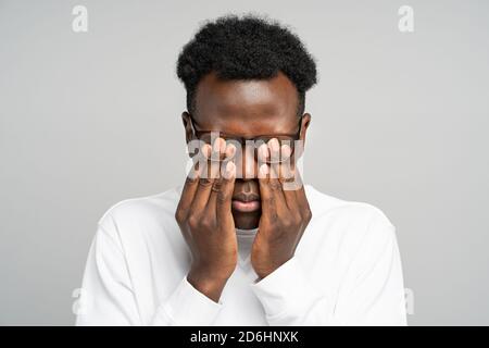 Close up of sleepy young afro-american man in glasses rubbing his eyes, feels tired after working on laptop, isolated on grey background. Overwork, ti Stock Photo