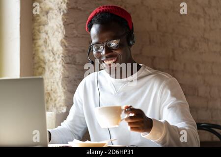 Happy millennial Black man in headphones wear red hat, enjoying watching educational webinar on laptop, remotely online work in cafe, holding cup of c Stock Photo