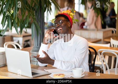 Happy millennial Afro-American man wear red hat laughing, speaking with friend on mobile phone, enjoying watching educational webinar on laptop, remot Stock Photo