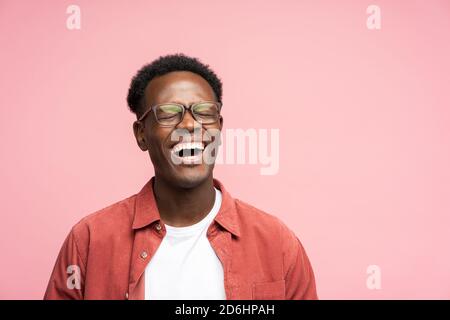 Laughing positive young Afro American man in red shirt in good mood, poses at camera with closed eyes. Overjoyed Black male in spectacles shows her br Stock Photo