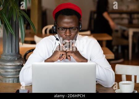 Focused millennial Black man in headphones wear red hat, watching educational webinar on laptop, remotely online work in cafe. Afro-American male list Stock Photo