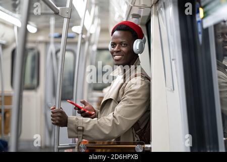Happy African American millennial man in red hat, trench coat stand in subway train, using mobile phone, listens to music with wireless headphones in Stock Photo