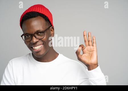 Cheerful playful young Afro American man wear red hat in good mood smiling broadly, showing okay gesture over studio grey background. Positive Black m Stock Photo