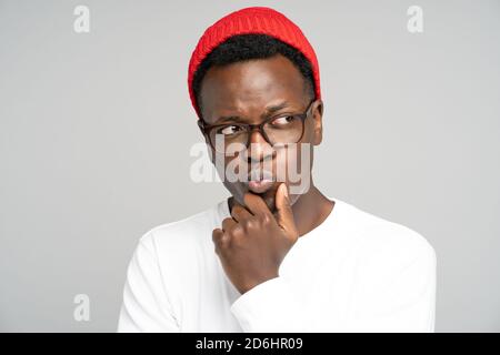 Studio portrait of unsure doubtful Black man in red hat wear glasses holds chin, looking right doubtfully, plans or reconsiders something, feeling hes Stock Photo